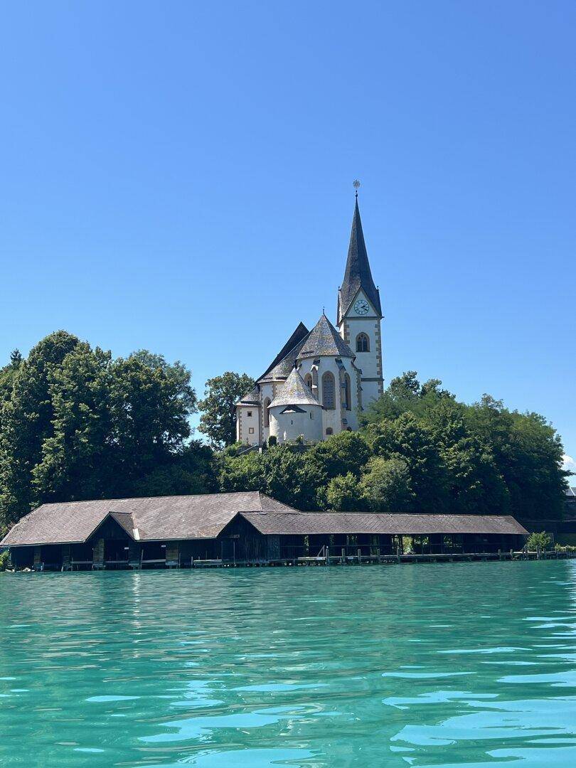 Historische Kirche auf einem Hügel mit traditionellen Bootshäusern am Ufer eines klaren Sees.