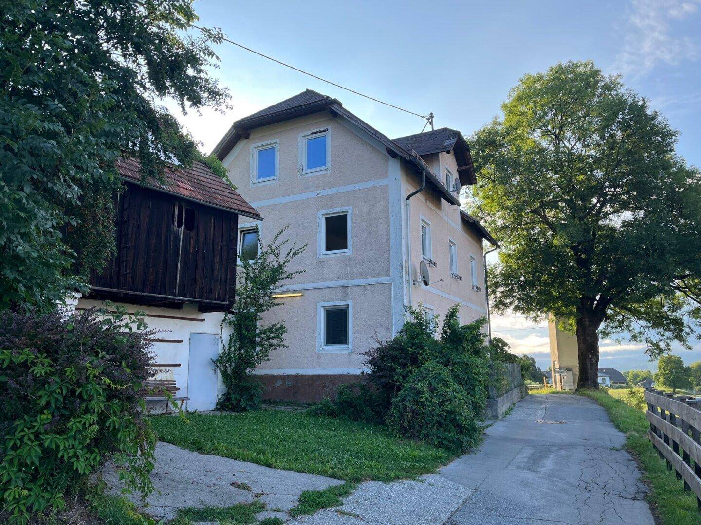 Traditionelles Landhaus mit Putzfassade, Holzanbau und großem Baum, umgeben von viel Grün.