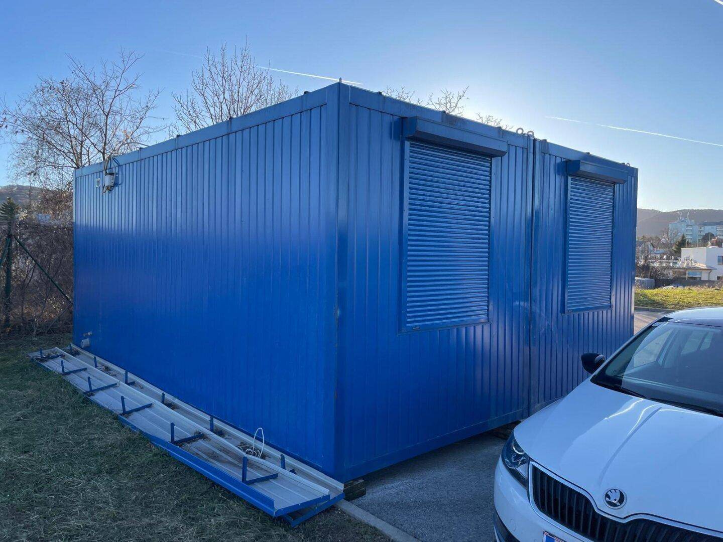 Ansicht des blauen Containergebäudes von der Seite mit geschlossenen Rollläden und angrenzendem Parkplatz.