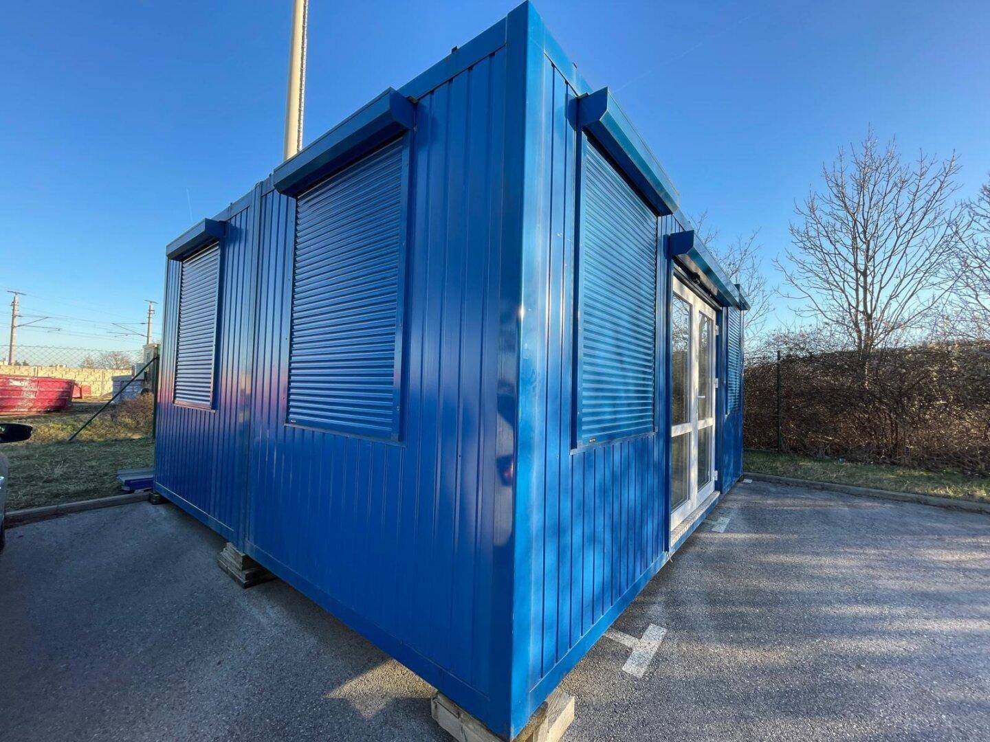 Blauer Containerbau von schräg vorne, mit Eingangstür und Fenstern, die mit blauen Rollläden versehen sind.