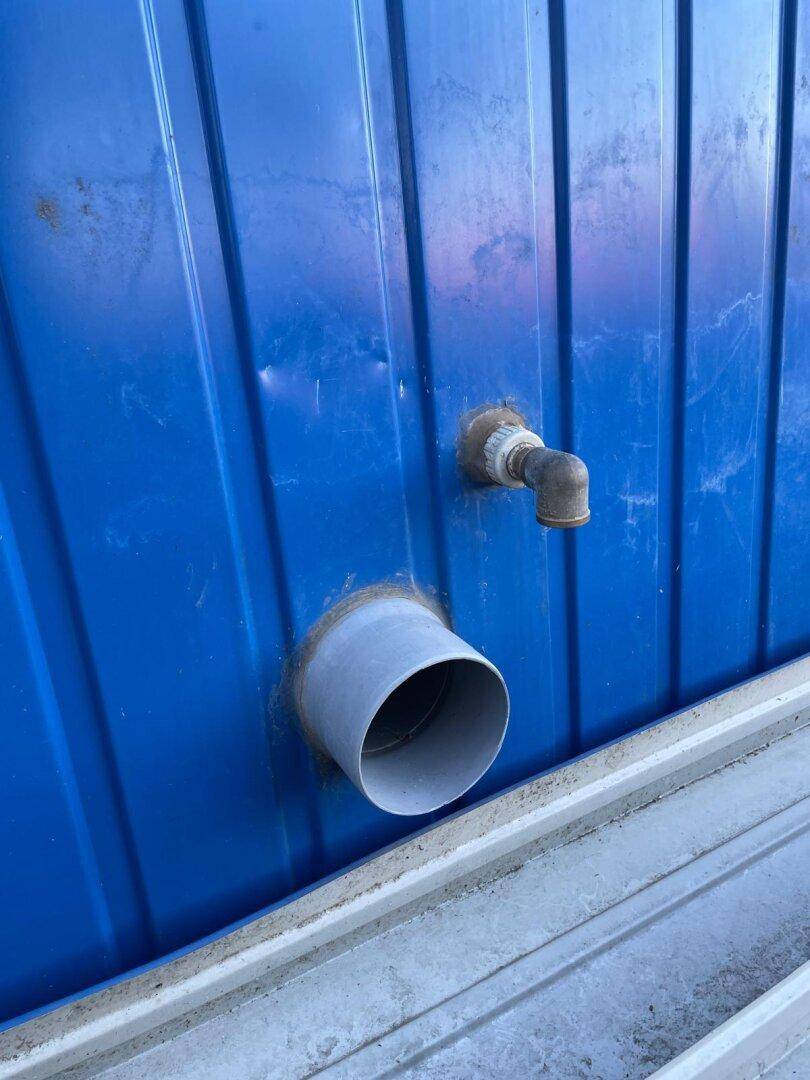 Nahaufnahme der blauen Metallfassade des Containers mit sichtbaren Rohranschlüssen und Entwässerung.