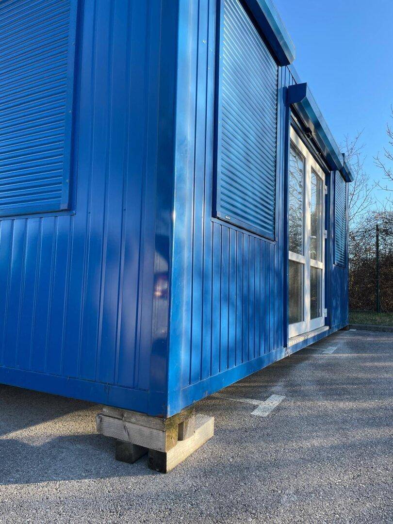 Detailansicht der blauen Containerfassade mit erhöhter Basis auf Holzblöcken und Fenster mit Rollladen.