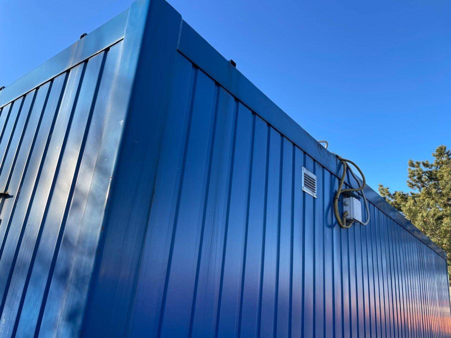 Detailansicht der oberen Ecke des blauen Containers mit Lüftungsgitter und Verkabelung an der Fassade.