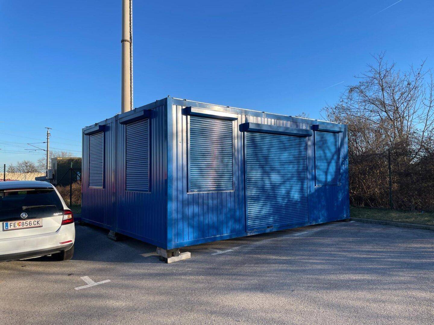 Gesamtansicht des blauen Containergebäudes mit Rollläden und einem geparkten Auto auf dem asphaltierten Bereich.
