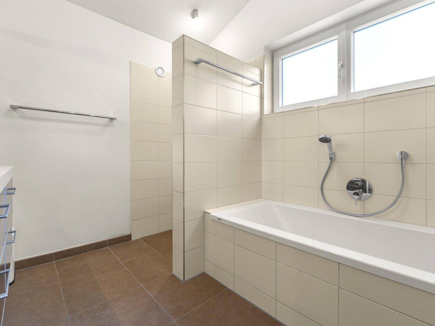 Badezimmer mit Badewanne, Dusche und Fenster, in neutralen Farbtönen gehalten.
