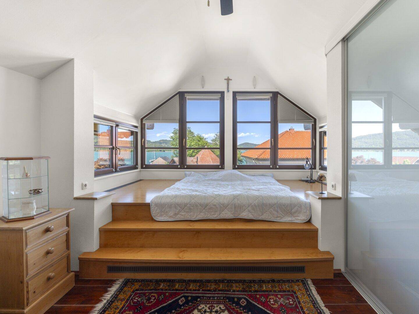 Schlafzimmer im Dachgeschoss mit erhöhtem Bettpodest und weitem Seeblick durch große Fenster.