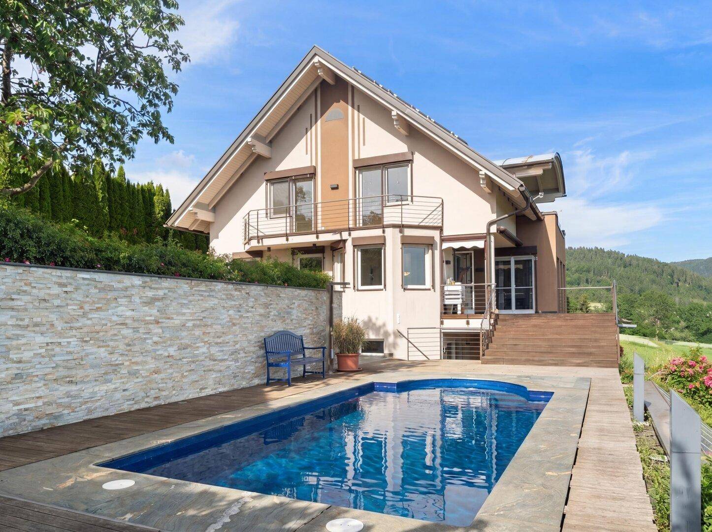 Haus mit blauem Pool und Holzdeck an einem sonnigen Tag, umgeben von grüner Natur.