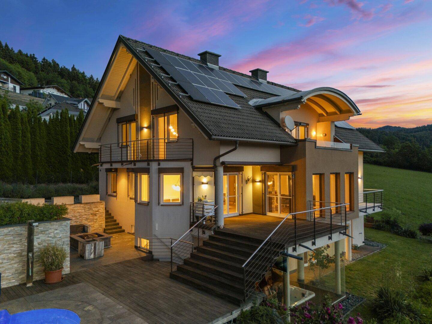 Haus mit beleuchtetem Pool, Solaranlagen und großem Holzdeck bei malerischem Sonnenuntergang.