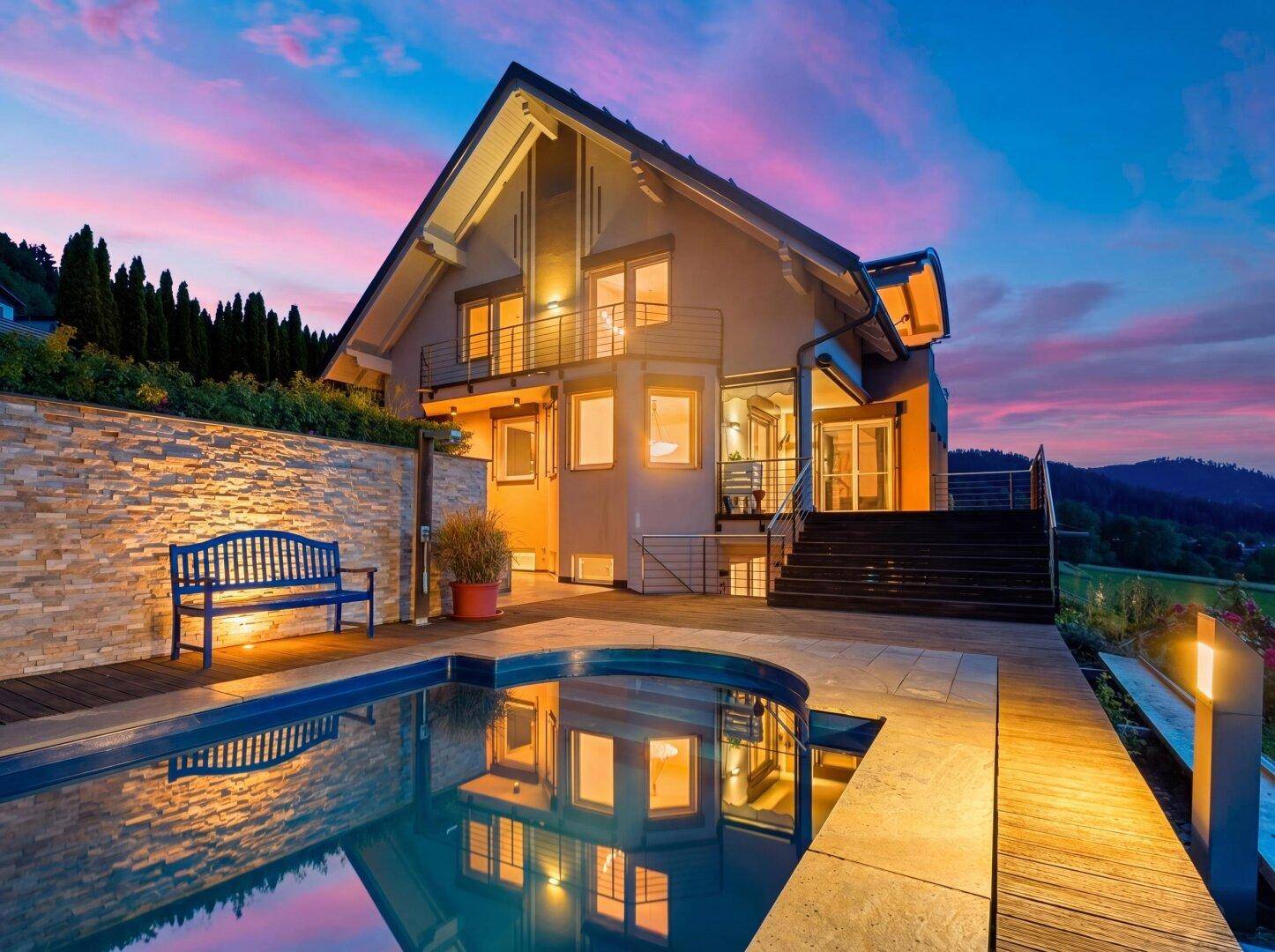 Haus mit beleuchtetem Pool und Holzdeck bei Sonnenuntergang, die Lichter spiegeln sich im Wasser.