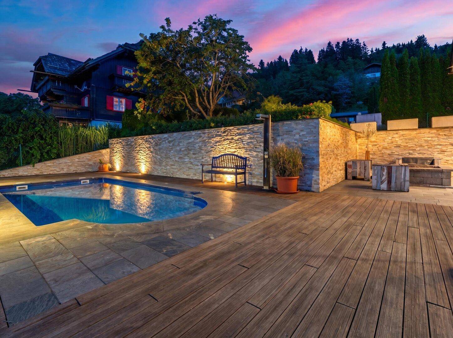 Beleuchteter Poolbereich mit Natursteinmauer und Holzdeck, umgeben von üppiger Vegetation am Abend.