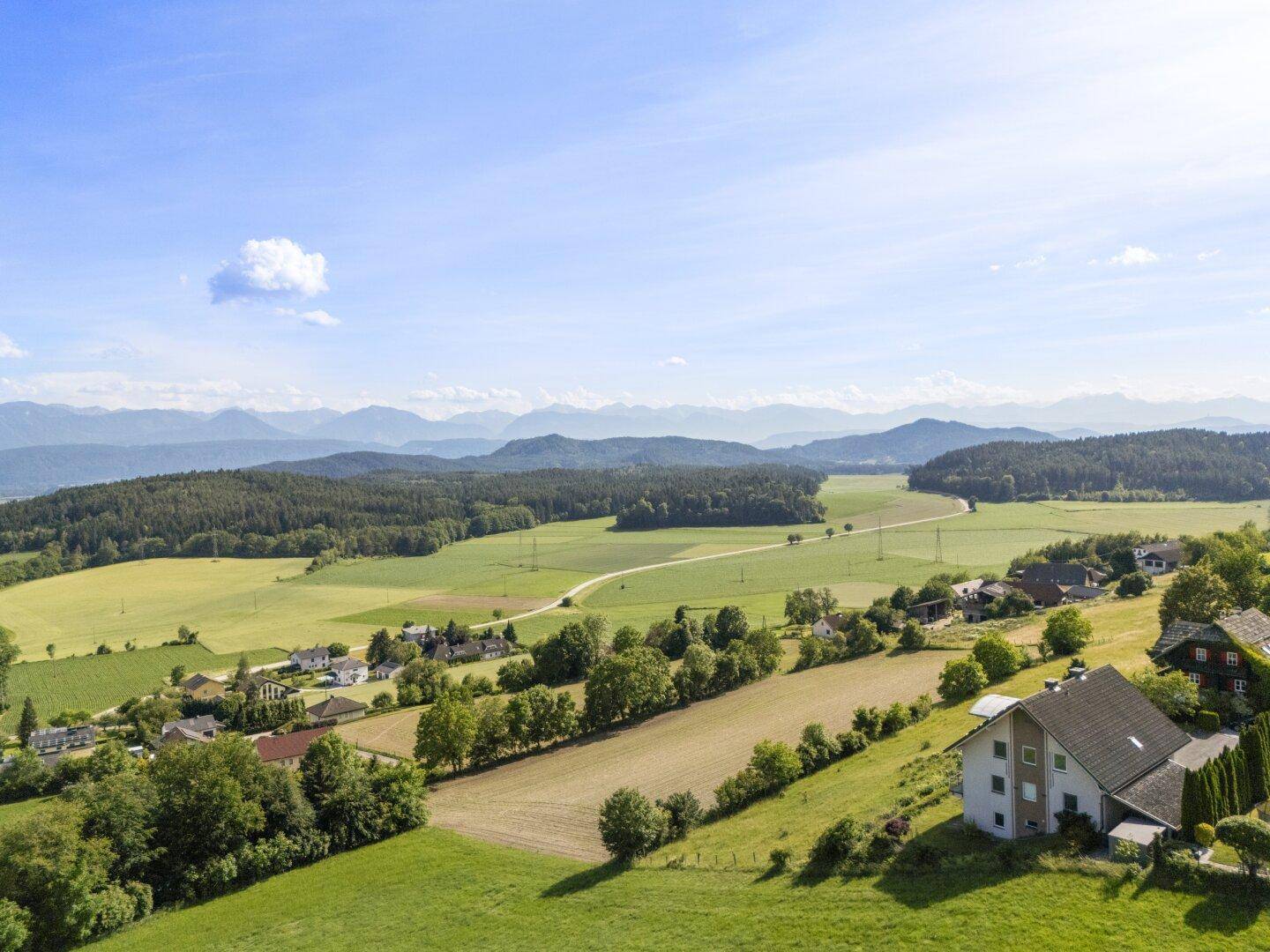 Weitläufige Landschaftsaufnahme mit grünen Feldern, Wäldern und verstreuten Häusern unter blauem Himmel.