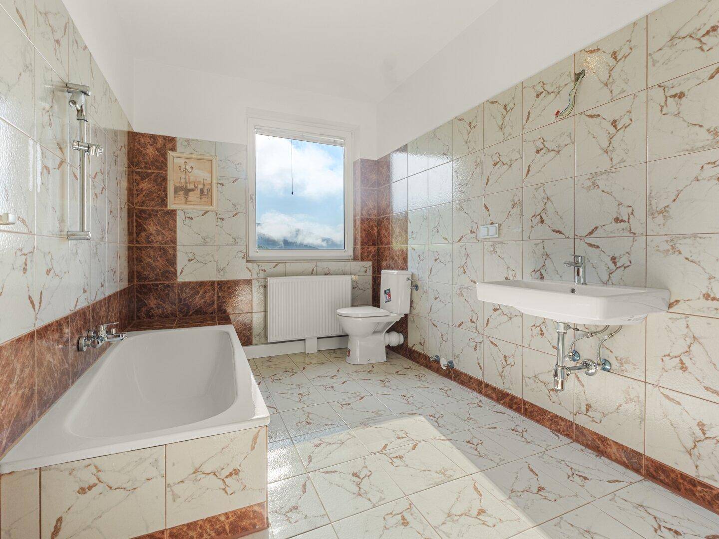 Geräumiges Badezimmer mit Badewanne, Fenster und gemusterten Fliesen in Braun- und Beigetönen.