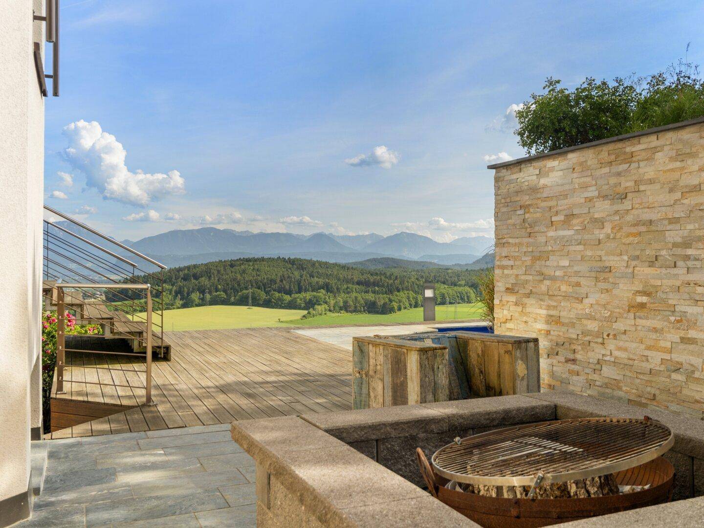 Außenbereich mit Grillplatz, Holzterrasse und Steinmauer, umgeben von Natur und Bergblick.
