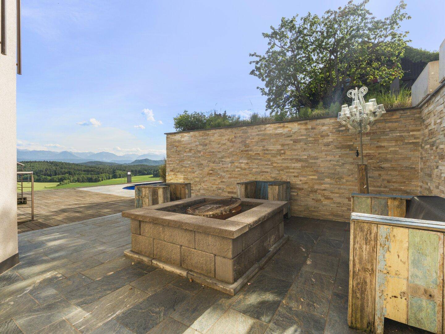 Gemütlicher Terrassenbereich mit Feuerstelle, Steinmauer und Blick auf die Landschaft.