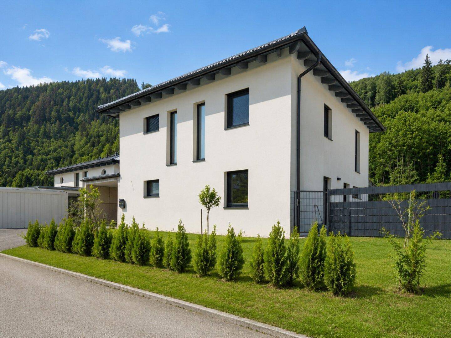 Moderne Hausfassade mit großen Fenstern und gepflegtem Vorgarten in grüner Umgebung.