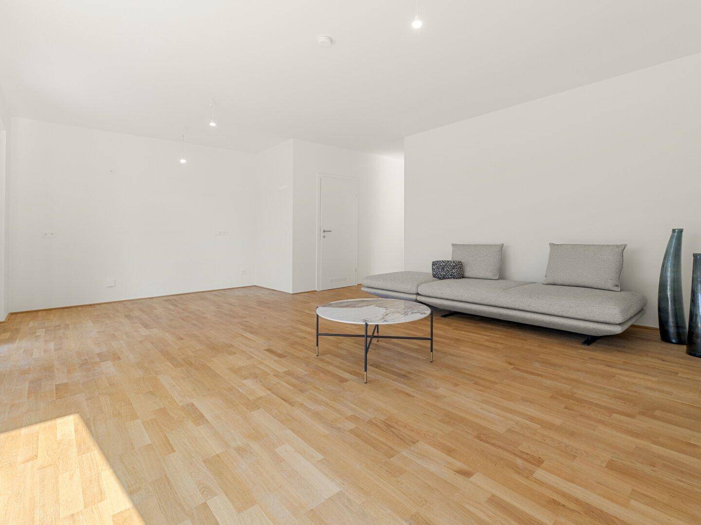 Geräumiges Wohnzimmer mit hellem Parkettboden, moderner Couch und viel Tageslicht.