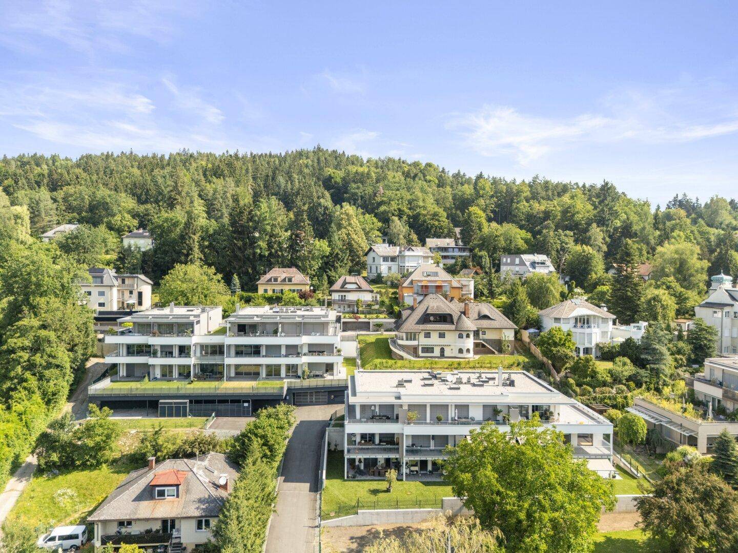 Luftaufnahme einer modernen Wohnsiedlung mit mehreren Gebäuden, umgeben von viel Grün und Hügellandschaft.