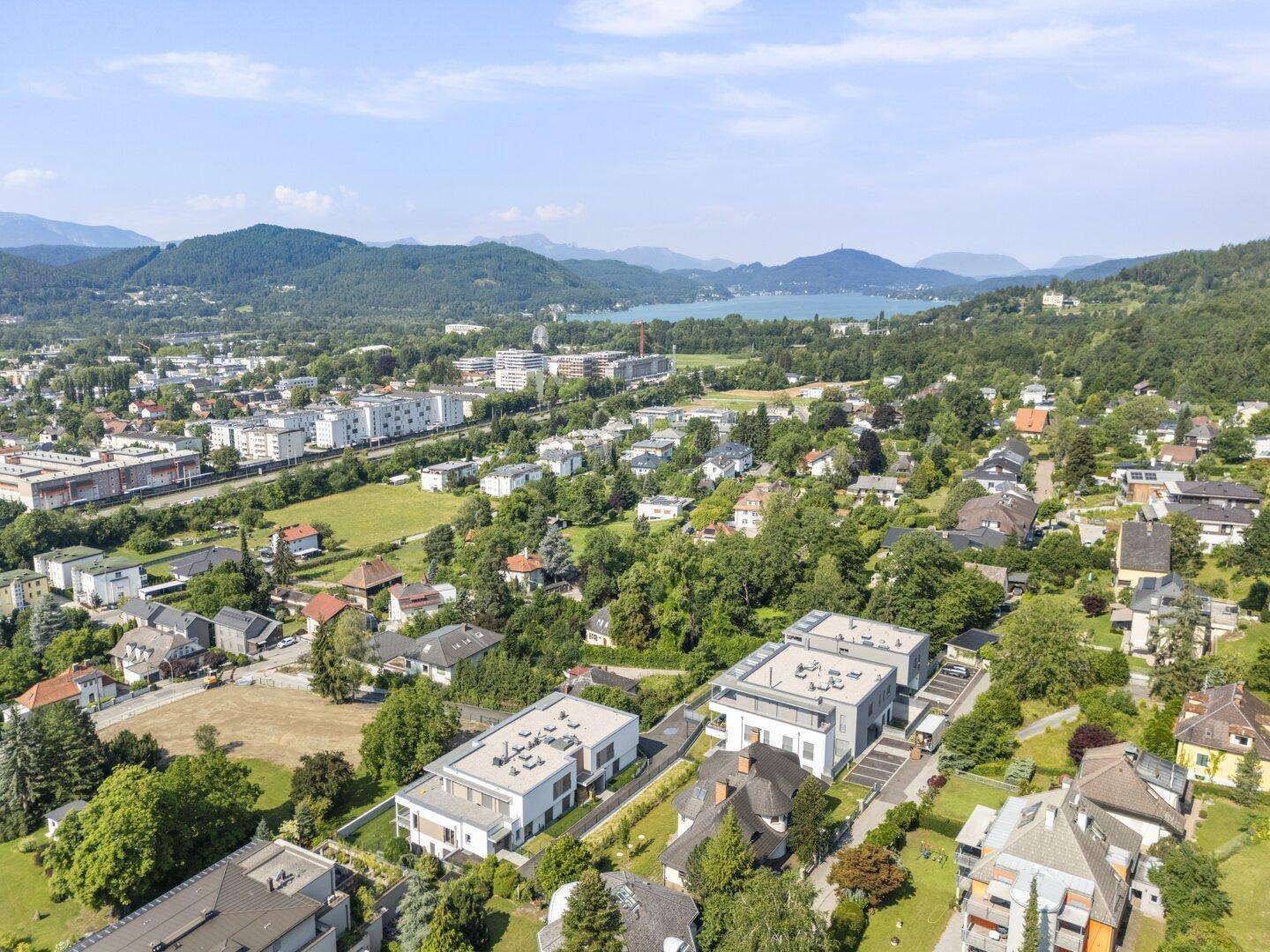 Weitläufige Luftaufnahme einer Stadt mit Wohngebäuden, umgeben von Natur, einem See und Bergen im Hintergrund.