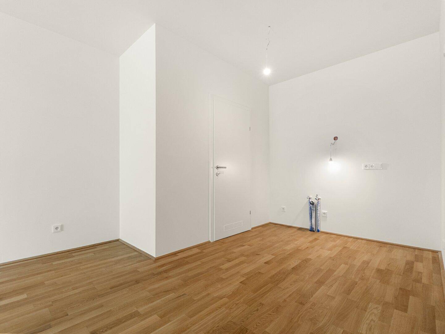 Leerer Raum mit Holzboden und vorbereiteten Anschlüssen für eine Küche, bereit zur Installation.