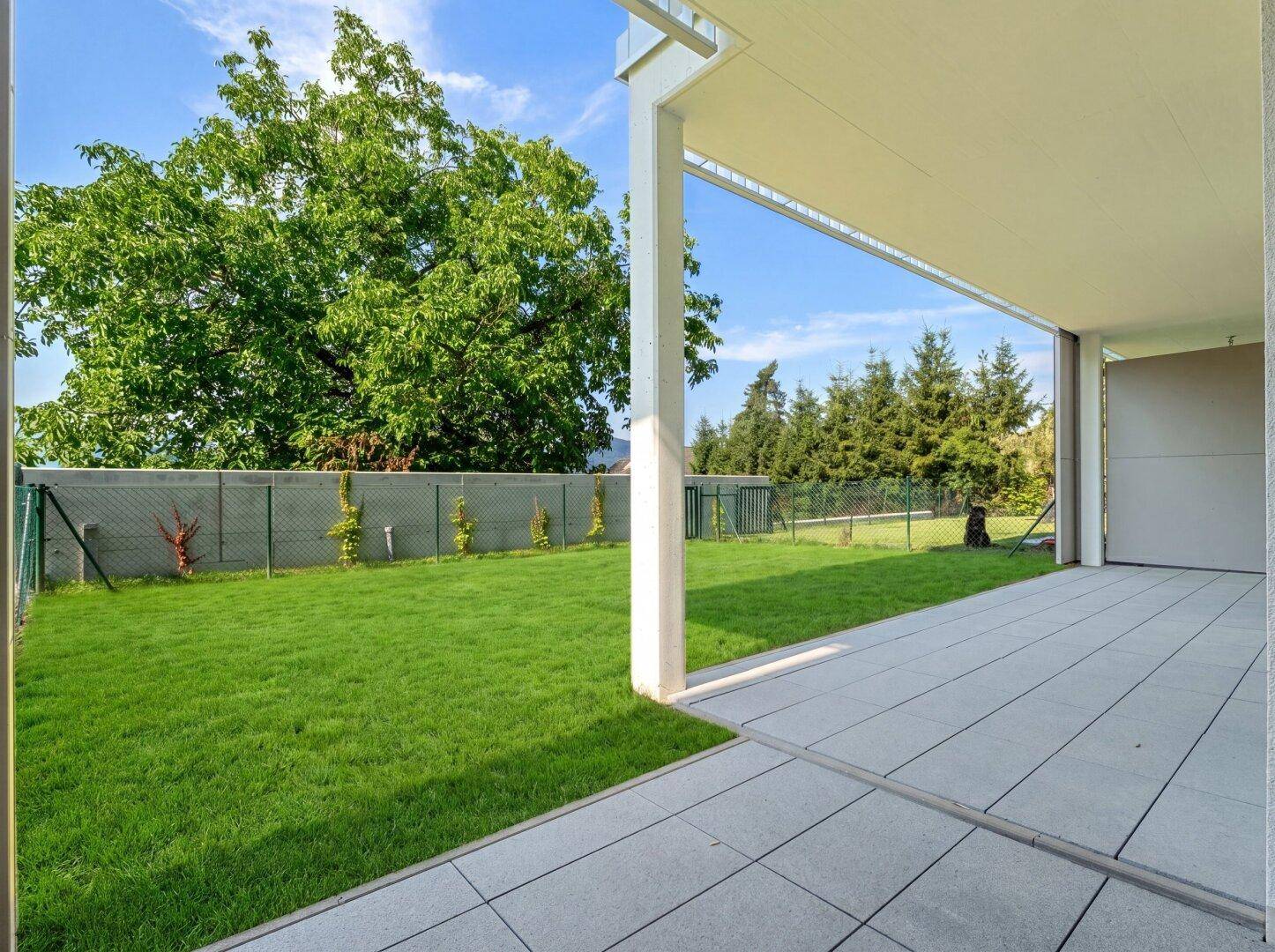 Geräumige geflieste Terrasse mit Überdachung und angrenzendem grünen Rasenbereich, umgeben von Bäumen.