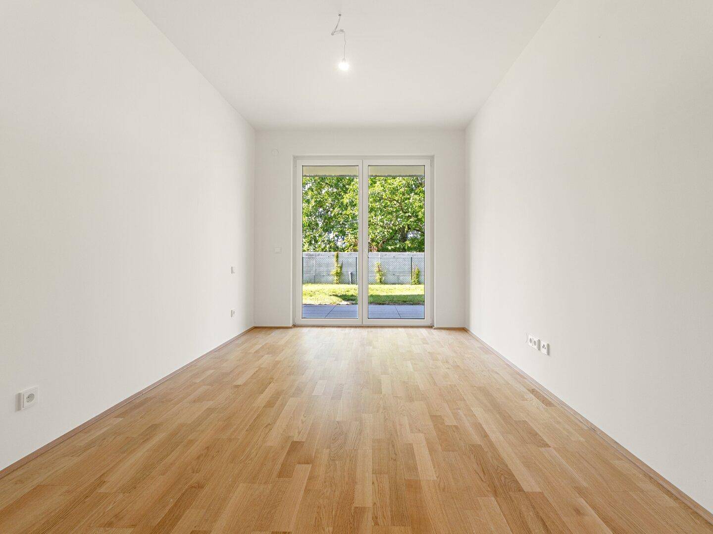 Heller, leerer Raum mit Holzboden und großen Schiebetüren, die zu einem grünen Außenbereich führen.