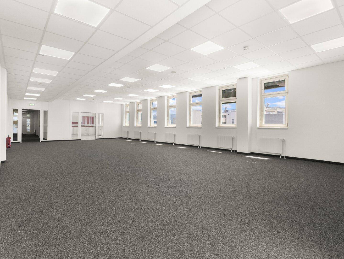 Weitläufiger Bürobereich mit Teppichboden, vielen Fenstern und Zugang zu einem Flur.