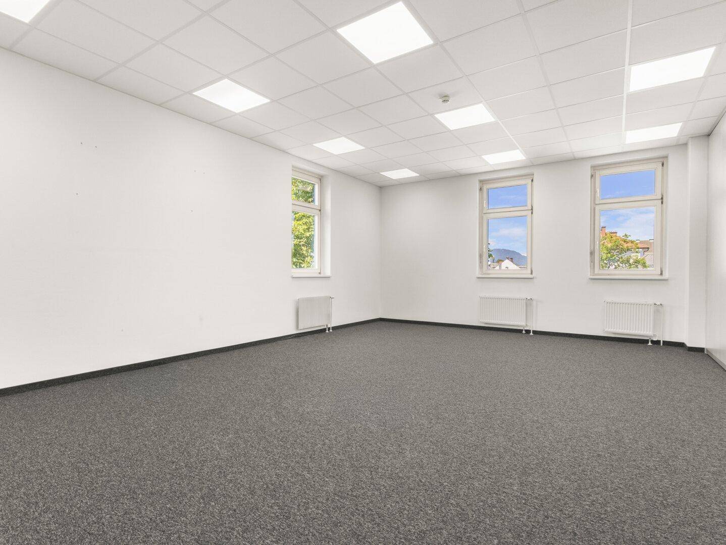 Heller Büroraum mit Teppichboden, weißen Wänden und mehreren Fenstern für Tageslicht.