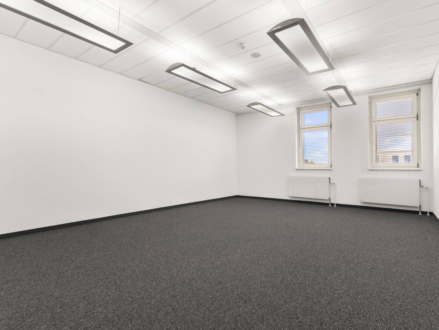 Kleinerer Büroraum mit Teppichboden, Jalousien an den Fenstern und modernen Hängelampen.