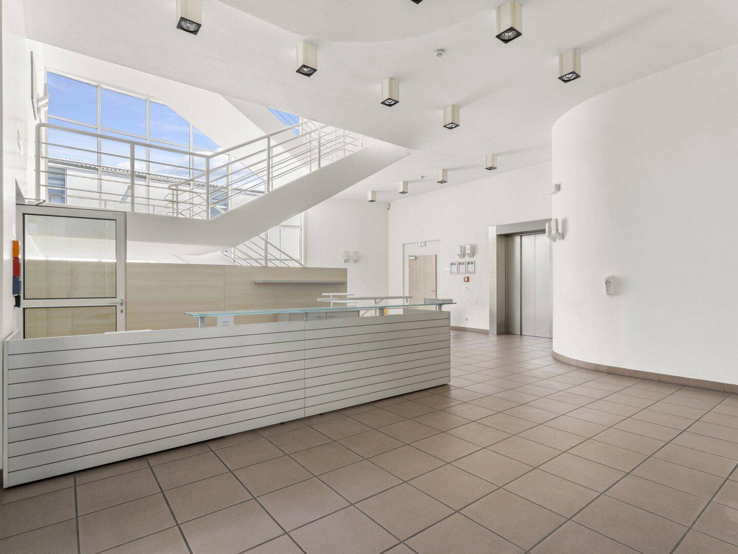Geräumiger Empfangsbereich mit Fliesenboden, Rezeption, großer Treppe und zwei Aufzügen.