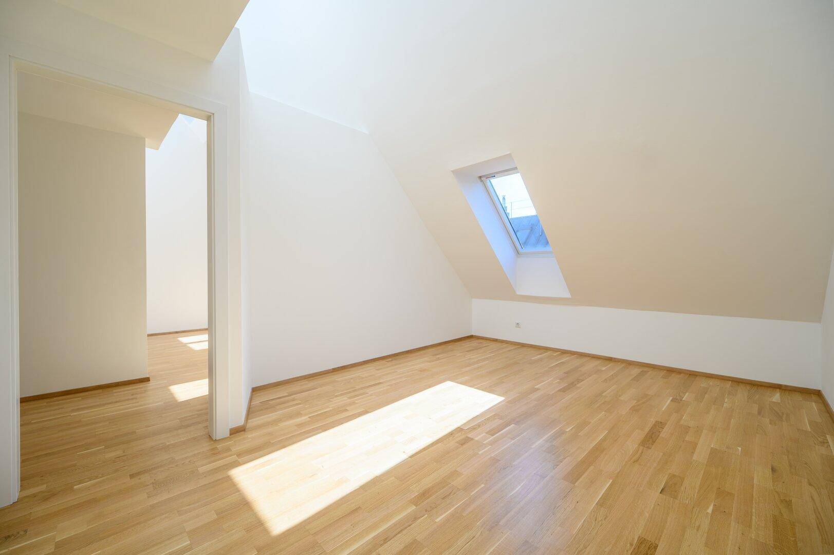 Lichtdurchflutetes Dachzimmer mit schrägen Wänden, Holzparkett und einem Dachfenster, das viel Helligkeit spendet.