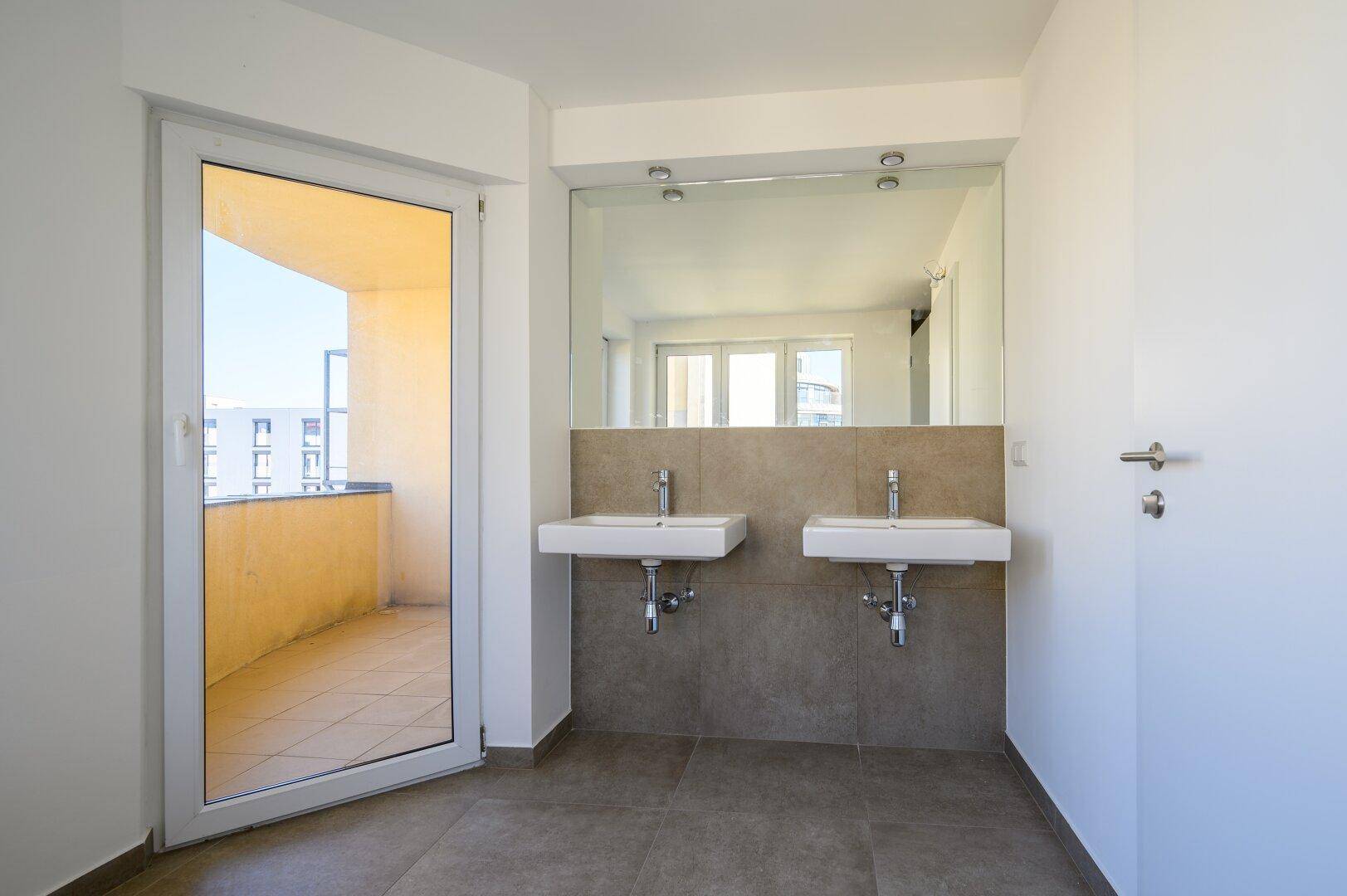 Modernes Badezimmer mit Doppelwaschbecken, großem Spiegel und direktem Zugang zu einem sonnigen Balkon.