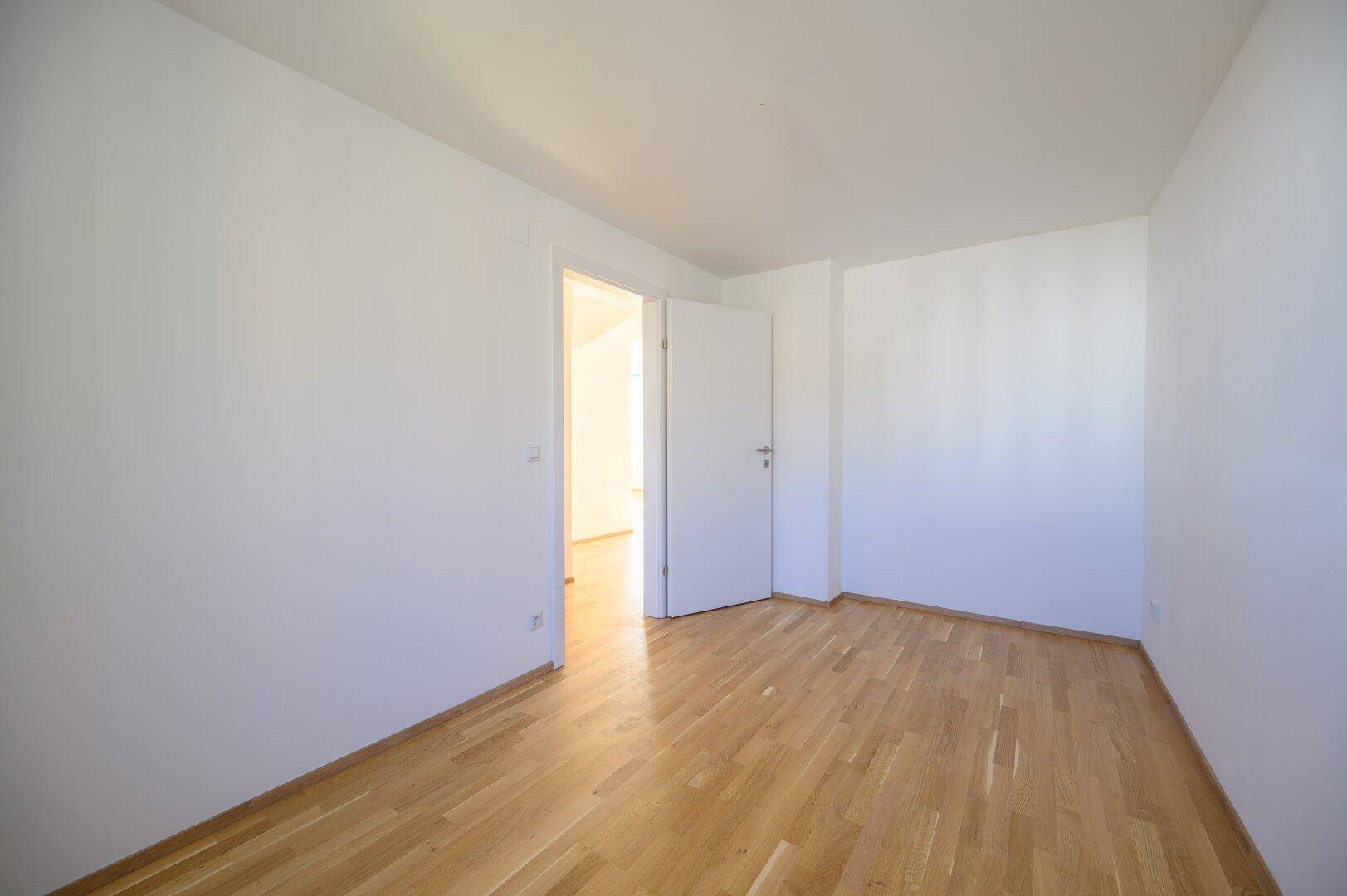 Helles, leeres Zimmer mit Holzparkettboden und weißen Wänden, bereit zur individuellen Gestaltung.