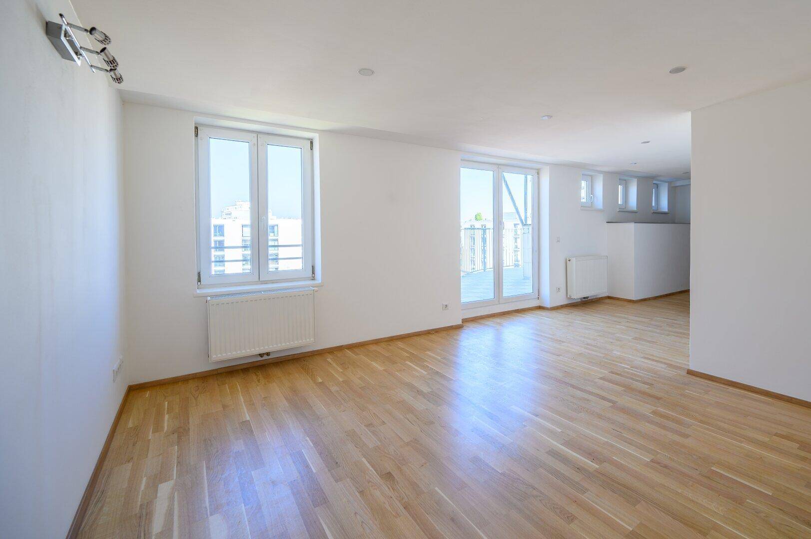 Heller Wohnbereich mit Holzparkett, großen Fenstern und Zugang zu einem Außenbereich, ausgestattet mit Heizkörpern.
