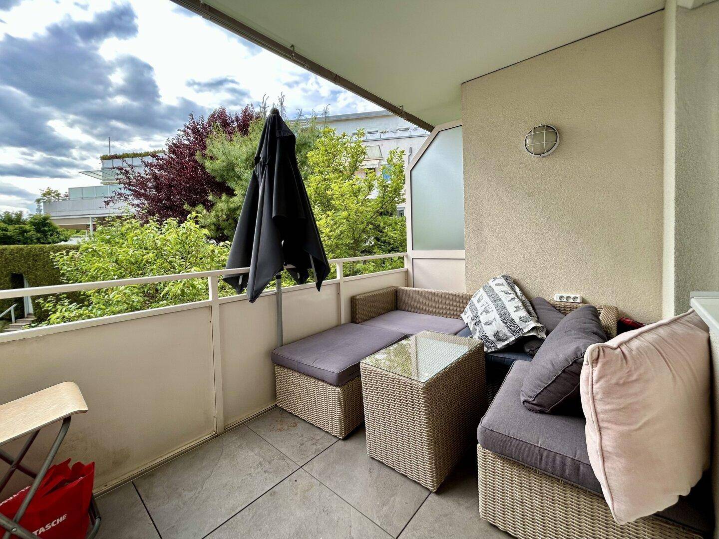 Gemütlicher Balkon mit Rattan-Lounge-Möbeln und Sonnenschirm, umgeben von Grün.