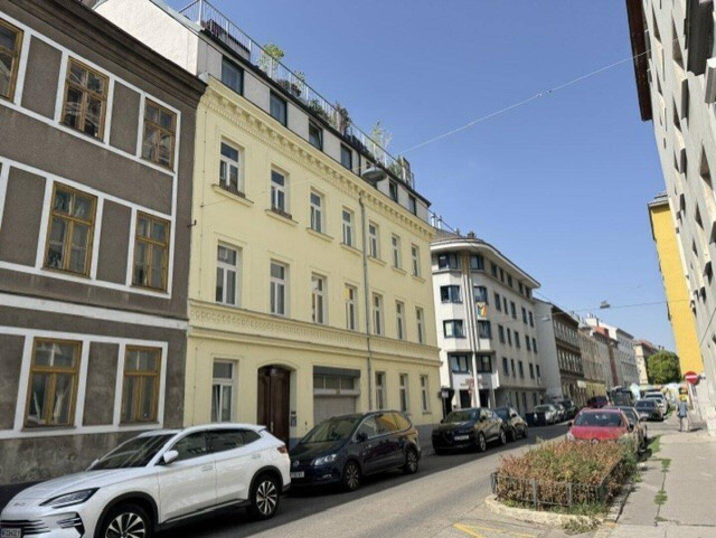Gebäude in einer belebten Stadtstraße mit parkenden Autos und umliegenden Häusern.