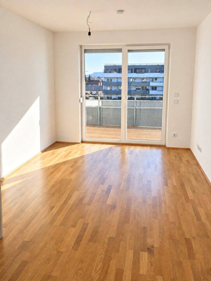 Geräumiges, helles Zimmer mit Holzboden und großem Fenster, das viel Tageslicht hereinlässt.