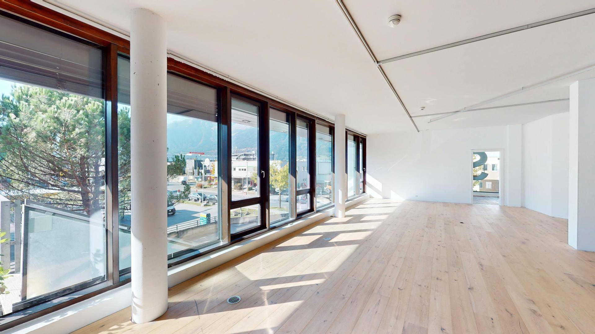 Langer, heller Raum mit durchgehender Fensterfront, Holzboden und weitem Blick nach draußen.
