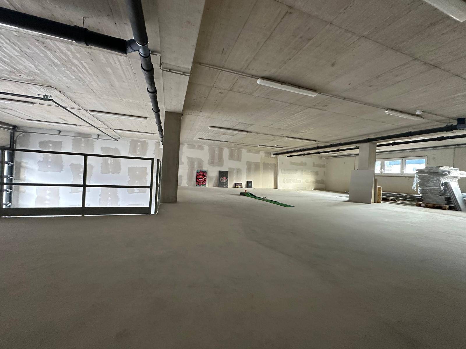 Weitläufiger Industrieraum mit Betonboden, sichtbaren Leitungen und unfertigen Wänden.