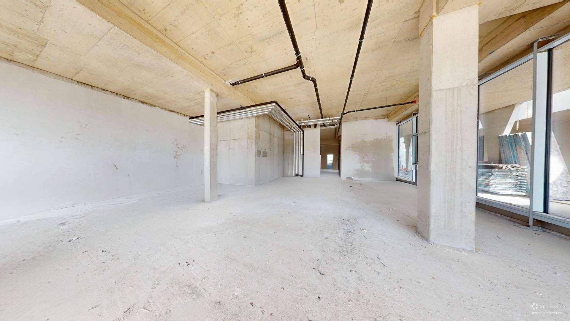 Geräumiger Rohbau mit Betonstützen, großen Fenstern und Blick auf die umliegende Baustelle.