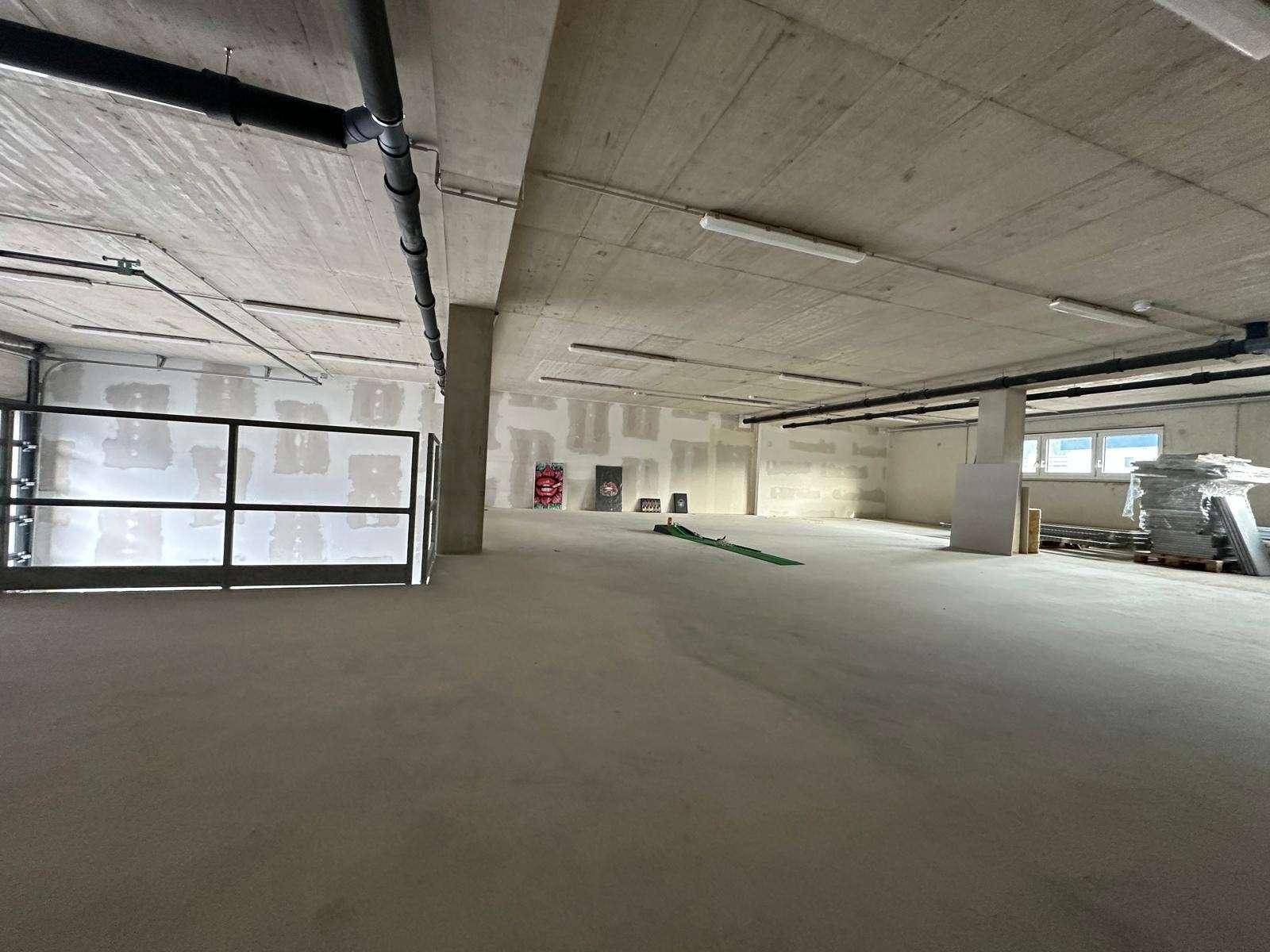 Weitläufiger Gewerberaum mit Sichtbetondecke, freiliegenden Installationen und teilweise verputzten Wänden.