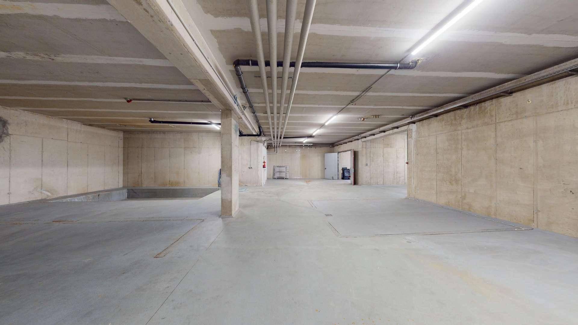 Geräumiger Kellerraum mit Betonwänden und -decken, sichtbaren Rohren und Beleuchtung.