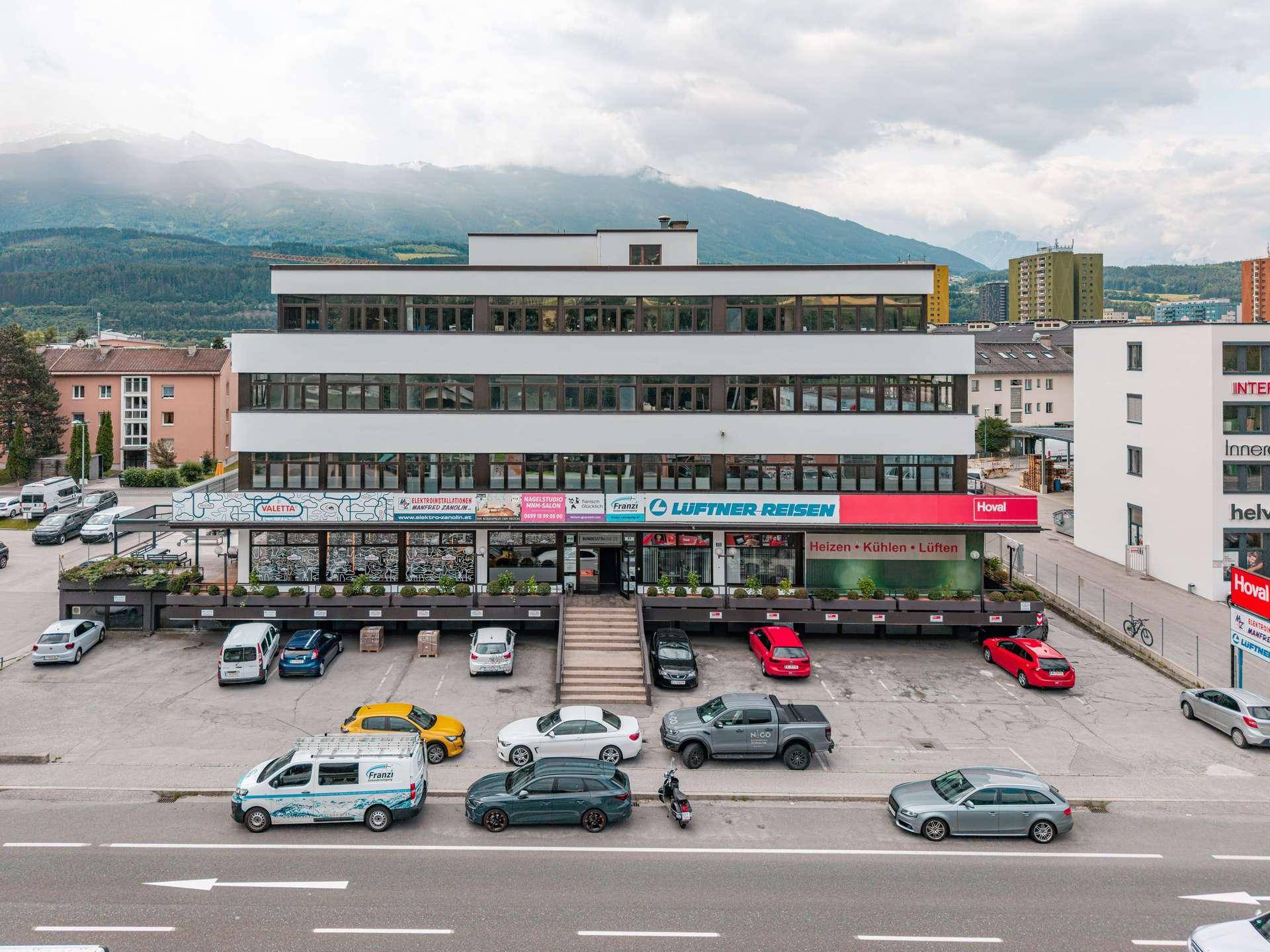 Modernes Geschäftsgebäude mit mehreren Etagen, umgeben von Parkplätzen und mit Bergblick im Hintergrund.