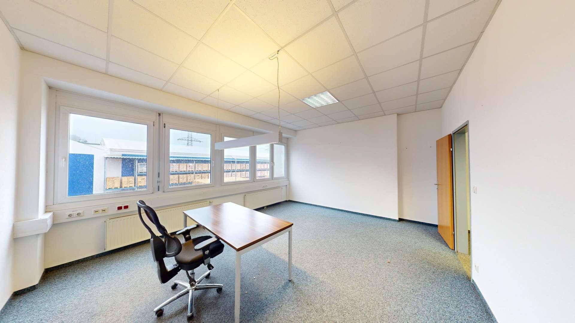 Helles und leeres Büro mit Teppichboden, einem einzelnen Schreibtisch und großen Fenstern.