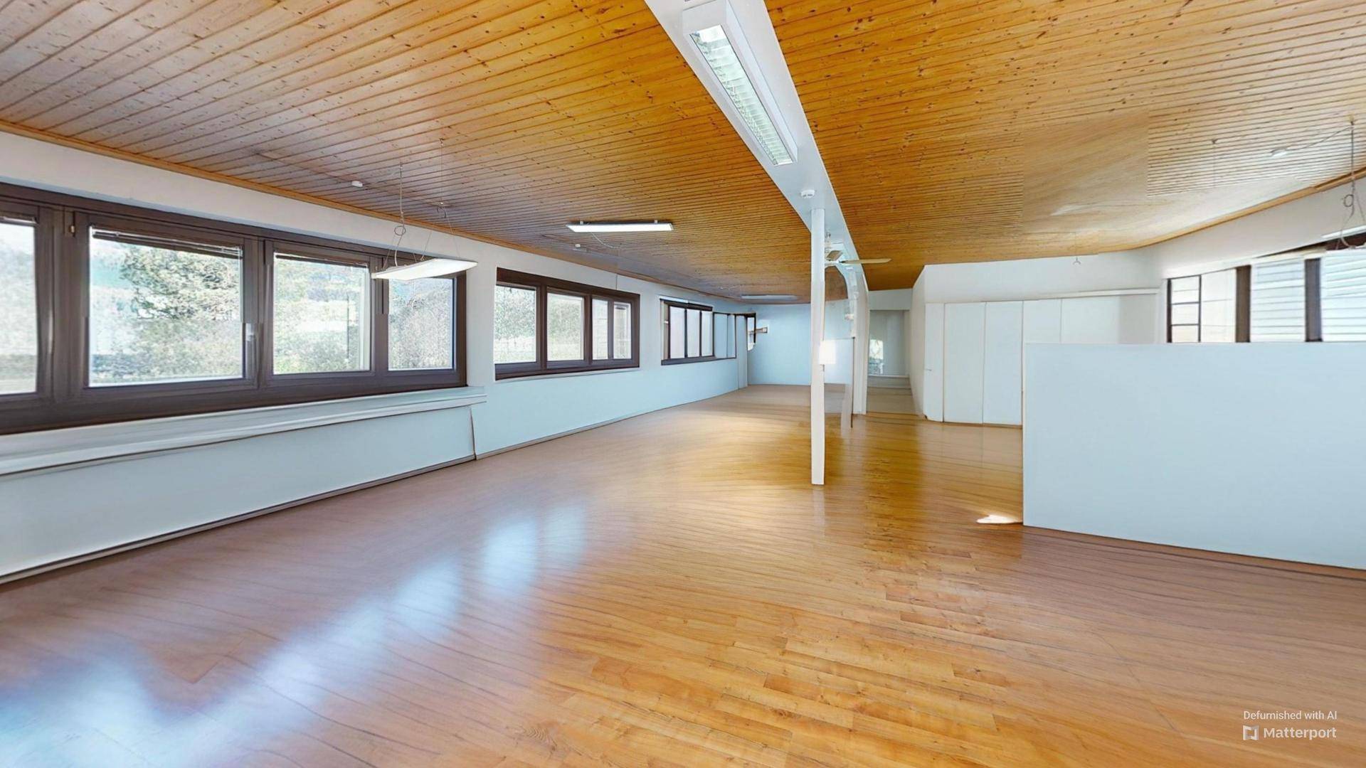 Großer, leerer Bürobereich mit Holzboden und Holzdecke, der viel natürliches Licht durch die Fenster erhält.