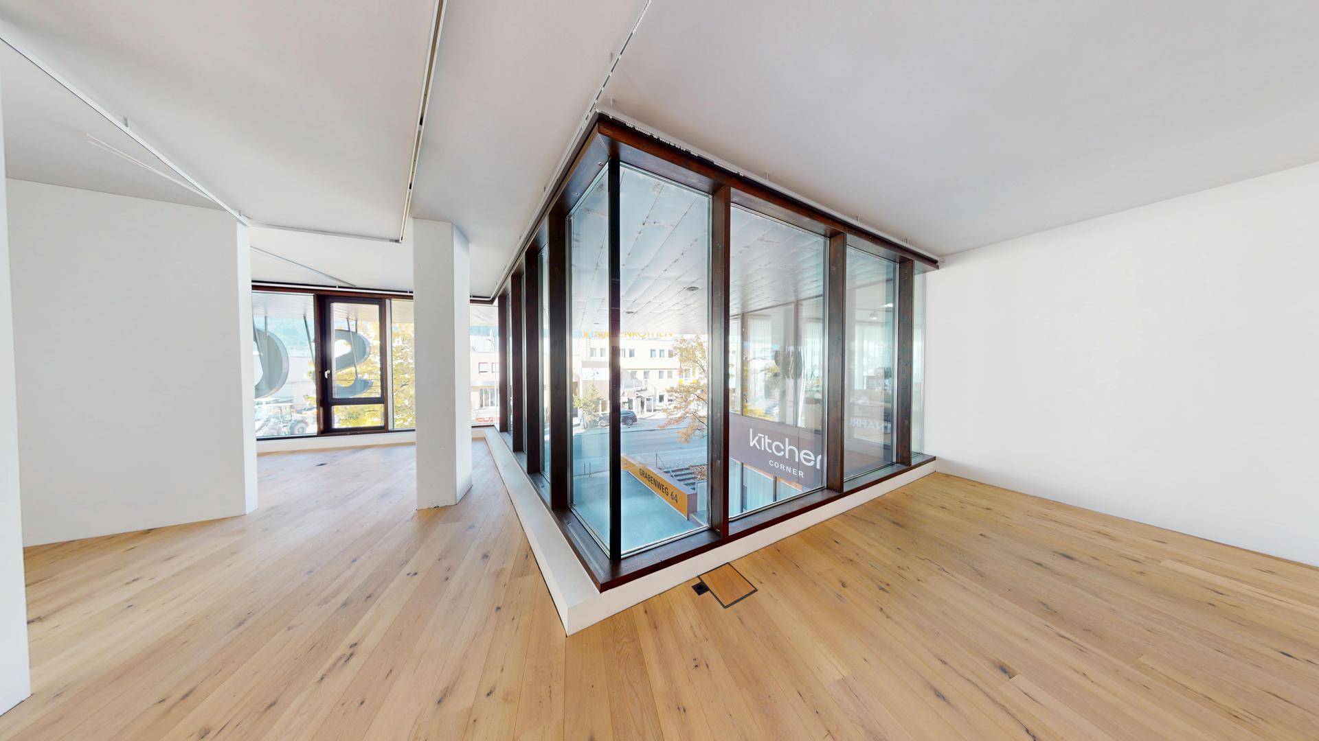 Leerer Innenraum mit Eckfenstern, Holzboden und Blick auf die Umgebung mit einem Küchengeschäft.