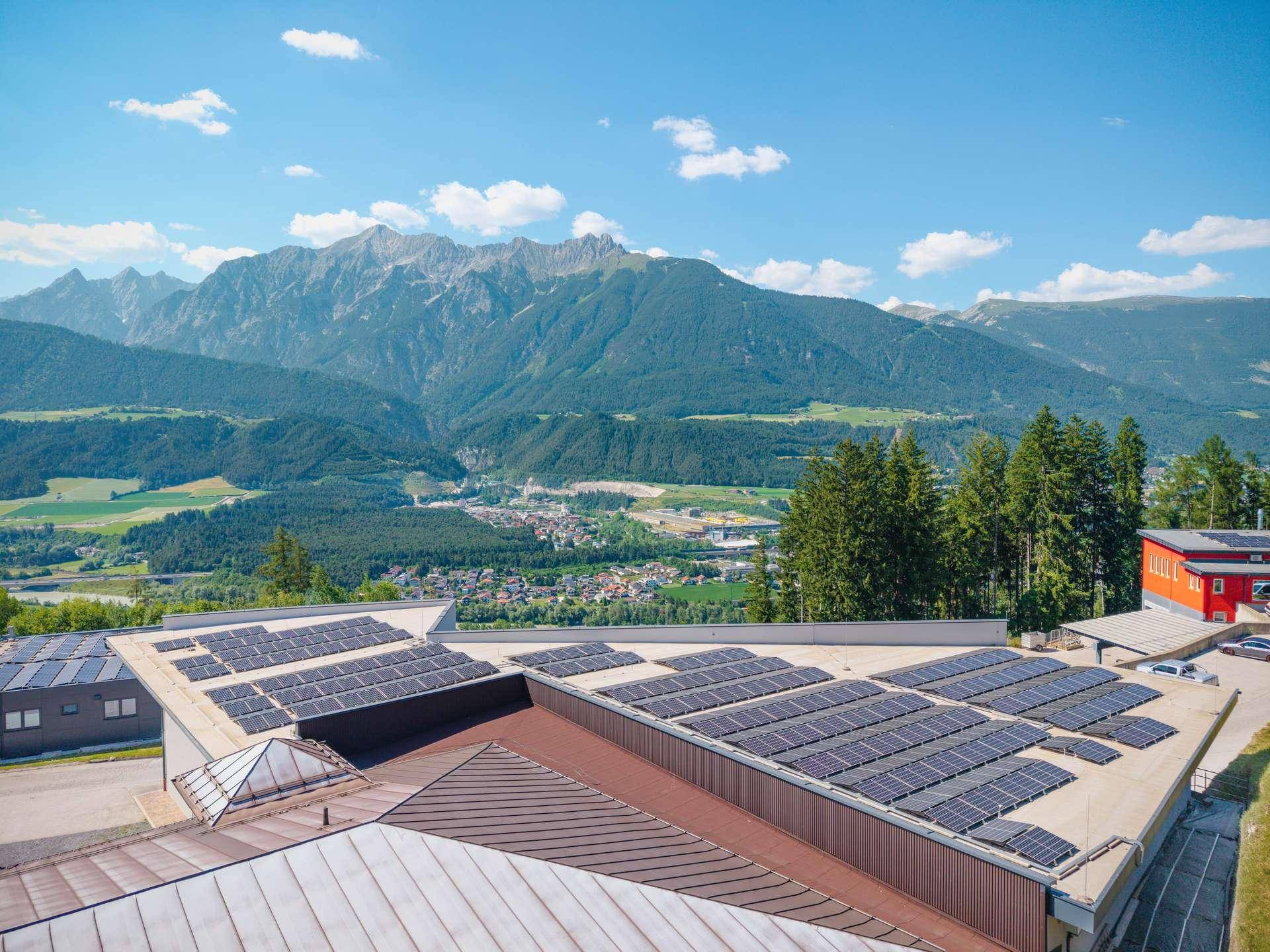 Luftaufnahme eines Gebäudes mit zahlreichen Photovoltaik-Modulen auf dem Dach und Bergblick.