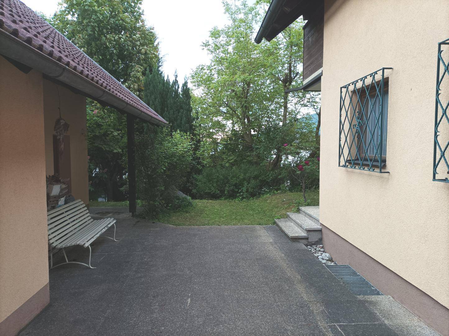 Gepflasterter Außenbereich neben dem Haus mit einer Bank und Zugang zum grünen Garten.