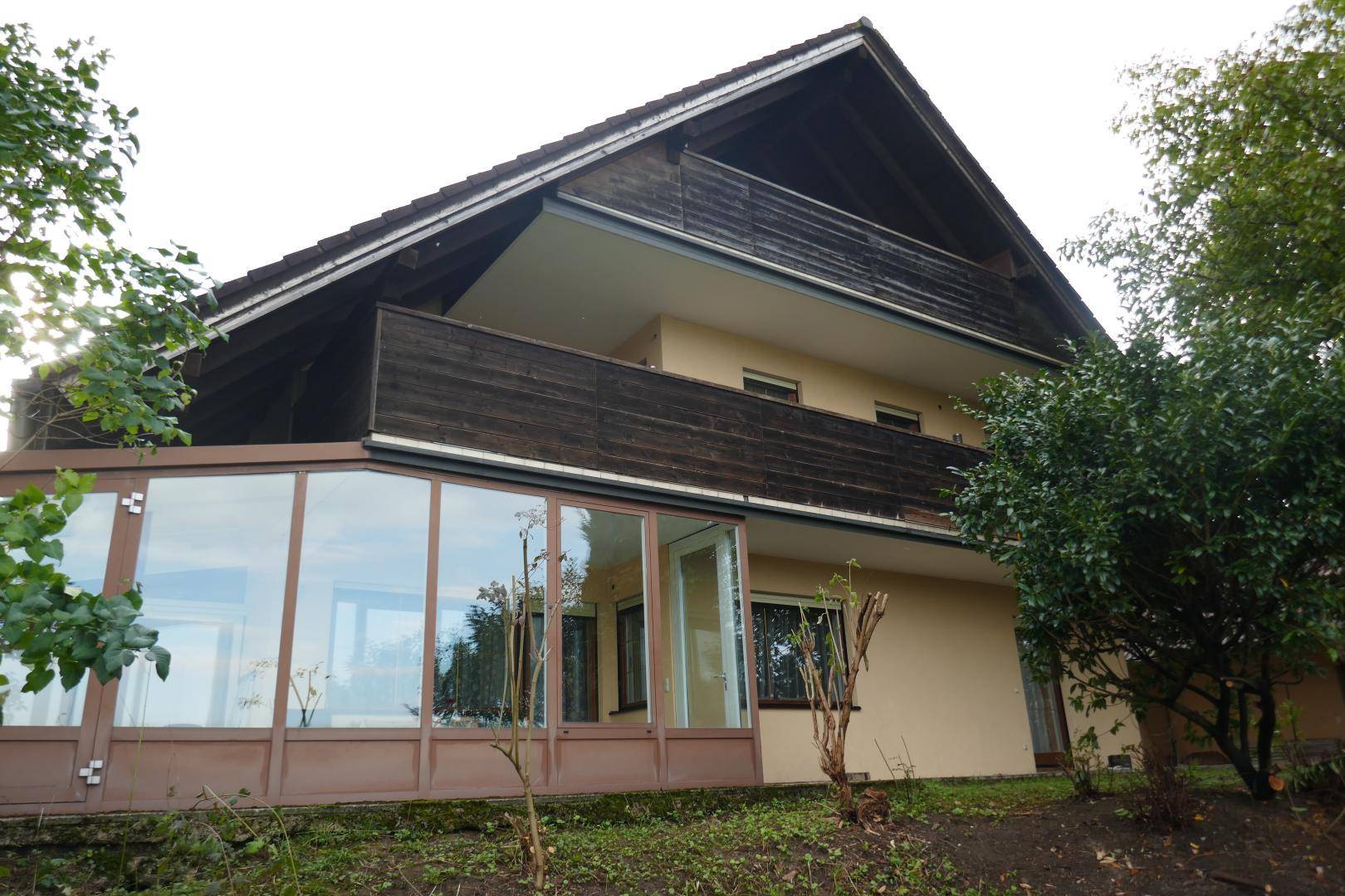 Rückansicht des Hauses mit Wintergarten im Erdgeschoss und einem Balkon im Obergeschoss, umgeben von Grün.