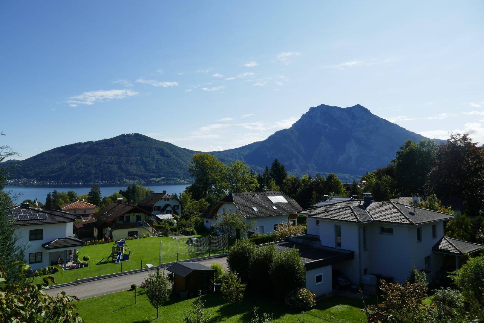 Panoramablick über eine Wohnsiedlung mit grünen Gärten, einem See und majestätischen Bergen im Hintergrund.
