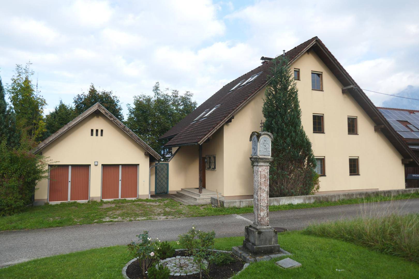 Traditionelles Wohnhaus mit Satteldach und separater Garage, umgeben von einem gepflegten Vorgarten mit Statue.
